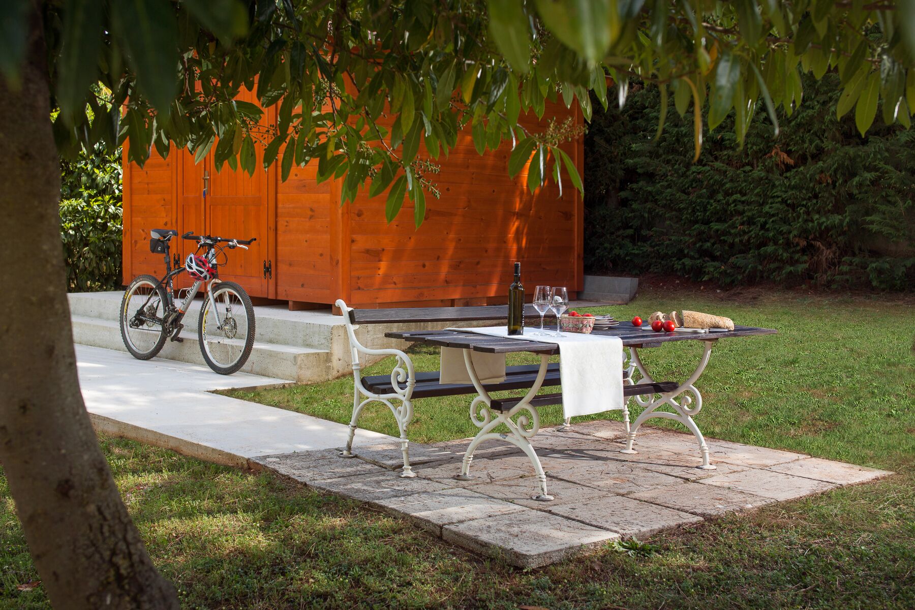 A picnic setup with a table, benches, and a bicycle in a garden area with a wooden structure in the background.