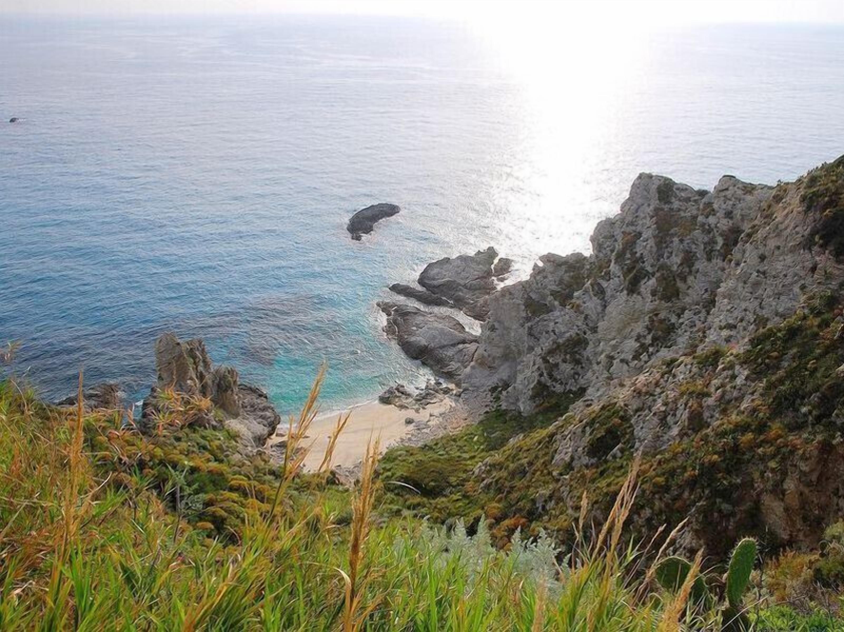 A coastal landscape with rocky cliffs, a beach, and a clear view of the sea.