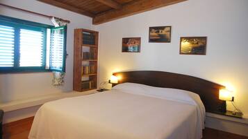 A bedroom with a large bed, wooden headboard, and two bedside lamps. There is a bookshelf and a window with blinds.