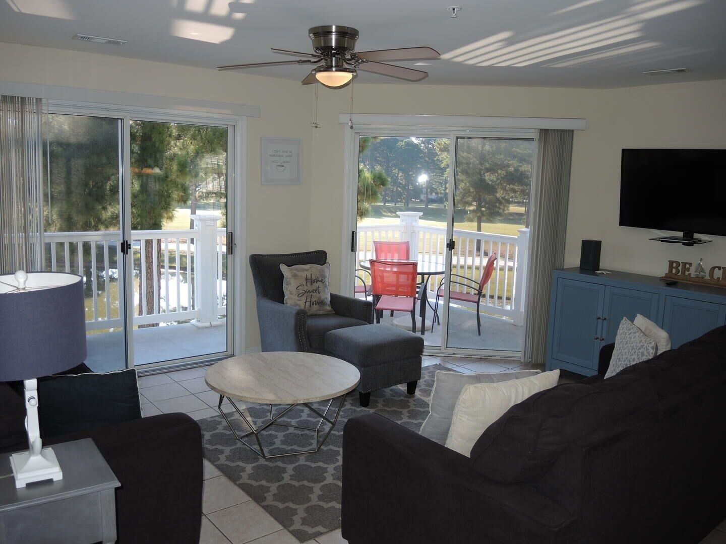 A living room with a sofa, armchair, and a small table. There is a ceiling fan, a TV on a cabinet, and a view of the outside through large sliding doors.