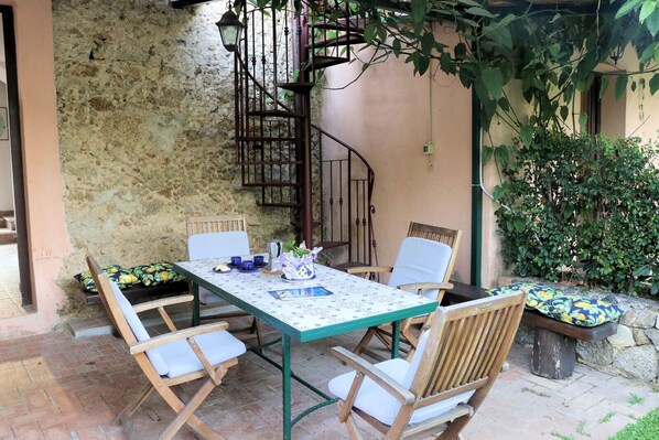 A patio with a table set for two, surrounded by wooden chairs. A spiral staircase is visible in the background, leading to an upper level.