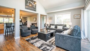 A spacious living room with a large sofa, a coffee table, and a fireplace. There is a kitchen area with bar stools and a dining table. The room has a high ceiling and large windows.