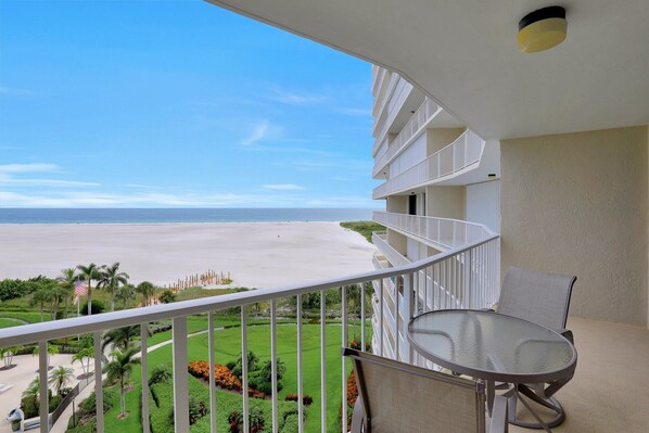 A balcony with a glass table and chairs overlooking a beach.