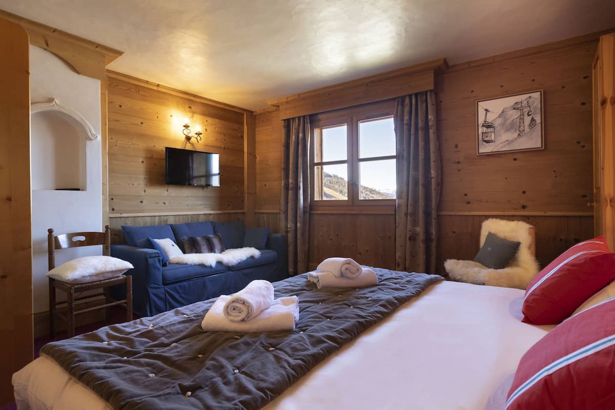 Una habitación de cabaña de madera con una cama, un sofá azul, una silla, un televisor montado en la pared y una ventana con vistas a las montañas.