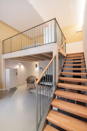 A modern interior with a wooden staircase leading to a loft area, a white refrigerator, and a wicker chair.