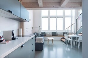 Eine moderne Küche mit integriertem Herd, ein Essbereich mit weißem Tisch und Stühlen sowie eine Sitzecke mit einer Bank und Kissen.