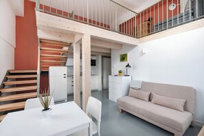 A modern interior with a staircase leading to a loft area, a dining table with a plant, a sofa, and a kitchen in the background.