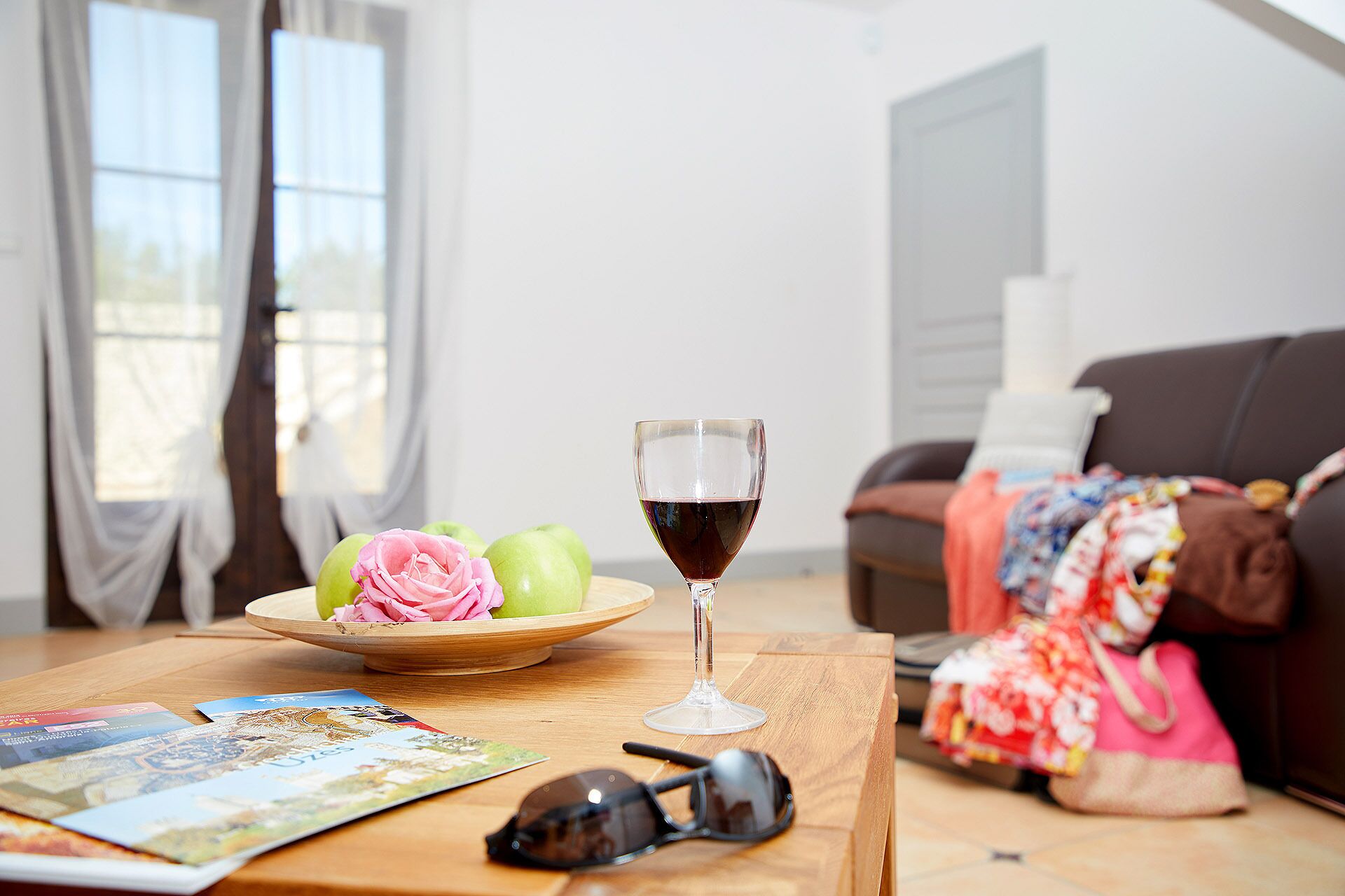 A wooden table with a glass of red wine, a bowl of green apples, and a pink rose.