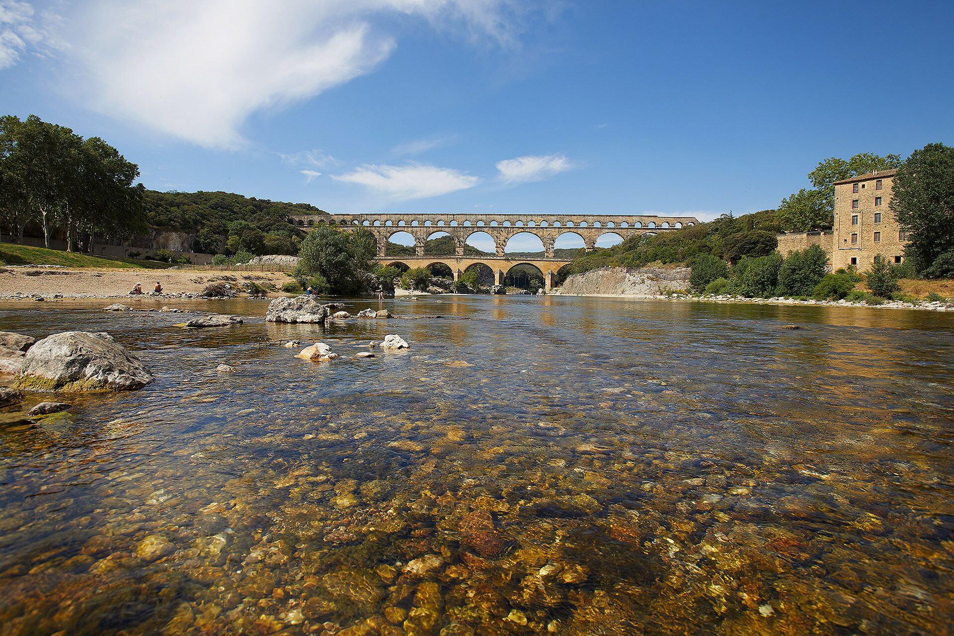 Un puente de piedra sobre un río cristalino con lecho rocoso, rodeado de vegetación y un edificio a la derecha.
