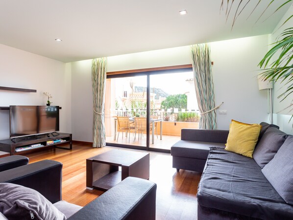 A modern living room with a flat-screen TV, sofa, coffee table, and a balcony with a view of the city.