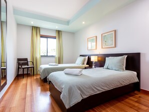 A hotel room with two beds, a chair, a window with curtains, and framed artwork on the wall.