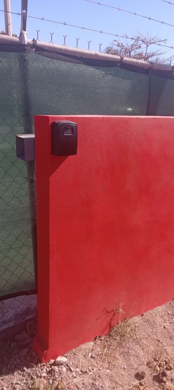 Una reja metálica roja con un picaporte negro, una cerca de malla verde y un cielo despejado al fondo.
