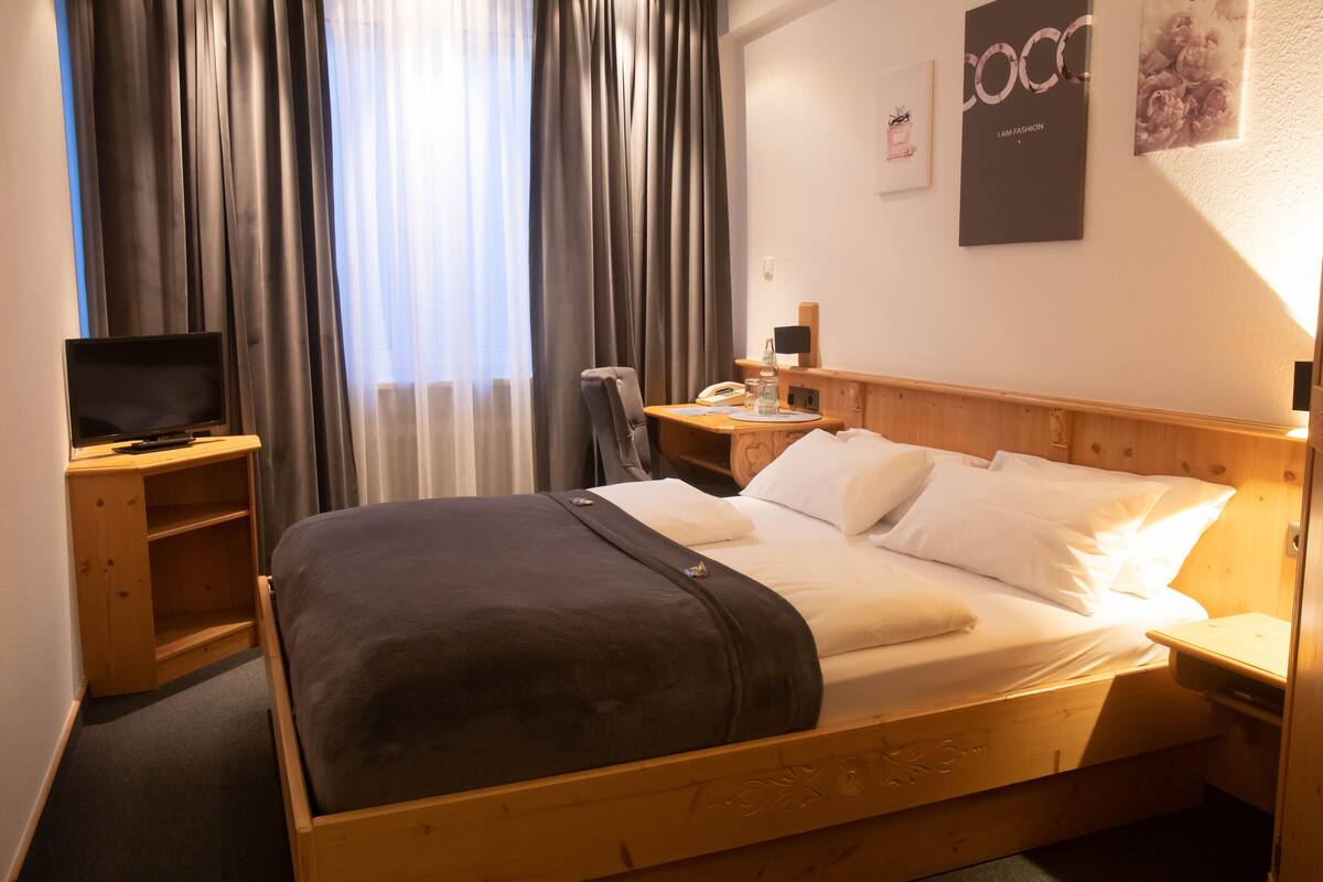 A wooden bed with a dark blanket, a bedside table with a lamp, a TV, and a wall-mounted phone.