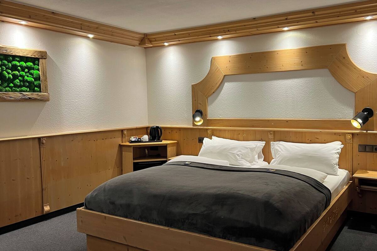 A neatly arranged hotel room with a wooden headboard, a bed with white linens, a bedside table with a lamp, and a small shelf with a kettle and cup.