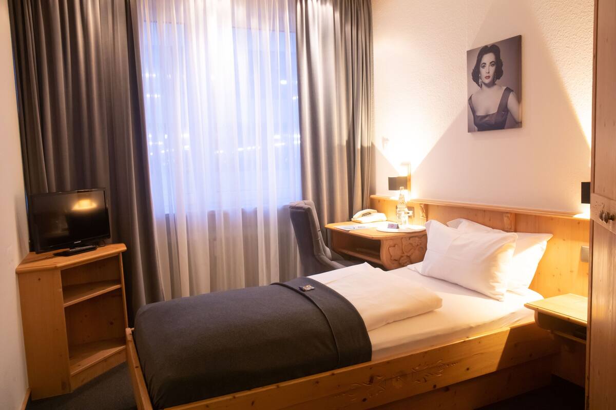 A hotel room with a bed, a TV, a desk with a lamp, a chair, and a window with curtains.