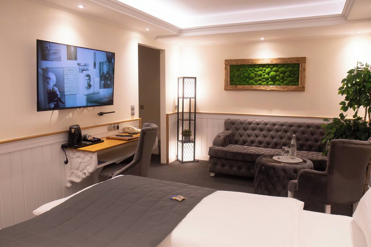 A modern hotel room with a large bed, a sofa, a TV mounted on the wall, and a small table with a plant.