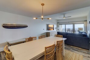 A hotel room with a dining area, a sofa, a ceiling fan, and a TV displaying a scenic view.