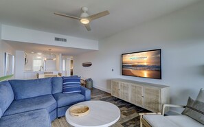 A modern living room with a blue sectional sofa, a round coffee table, a television mounted on the wall, and a ceiling fan.