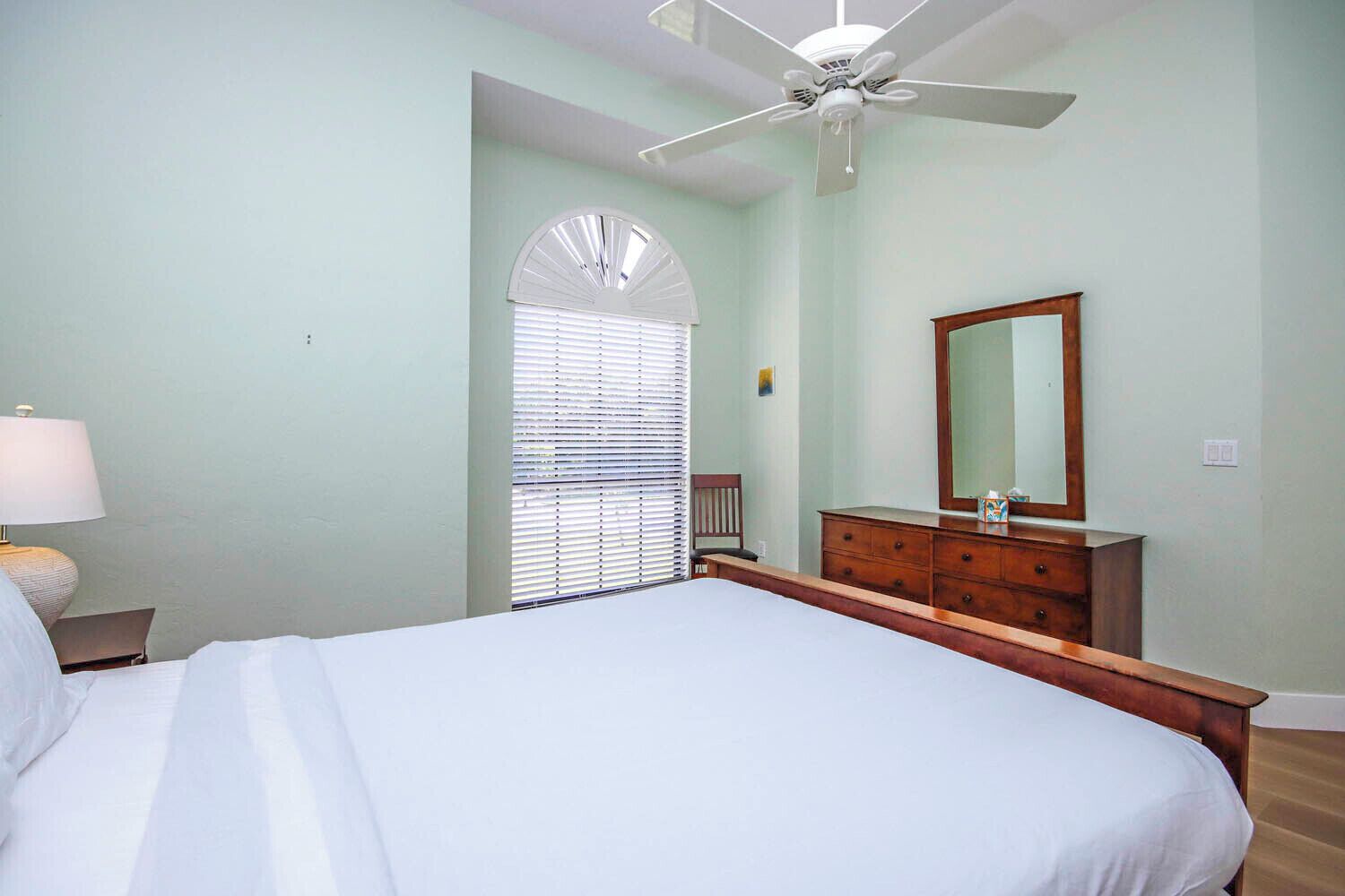 Une chambre à coucher comprenant un lit, une commode, un miroir, un ventilateur de plafond et une fenêtre avec des stores.