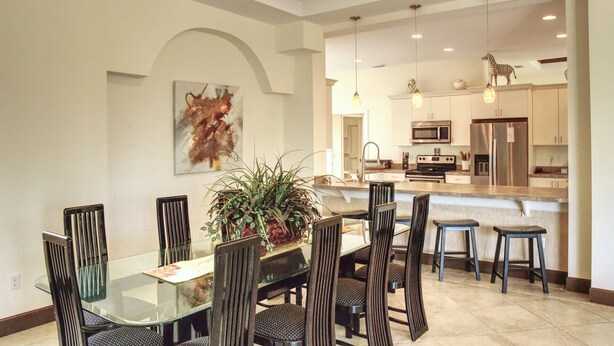 A dining area with a glass table and chairs, a potted plant, and a kitchen in the background. - Luxury Gulf Beach Living - Coronas Gris (Siesta Key)
