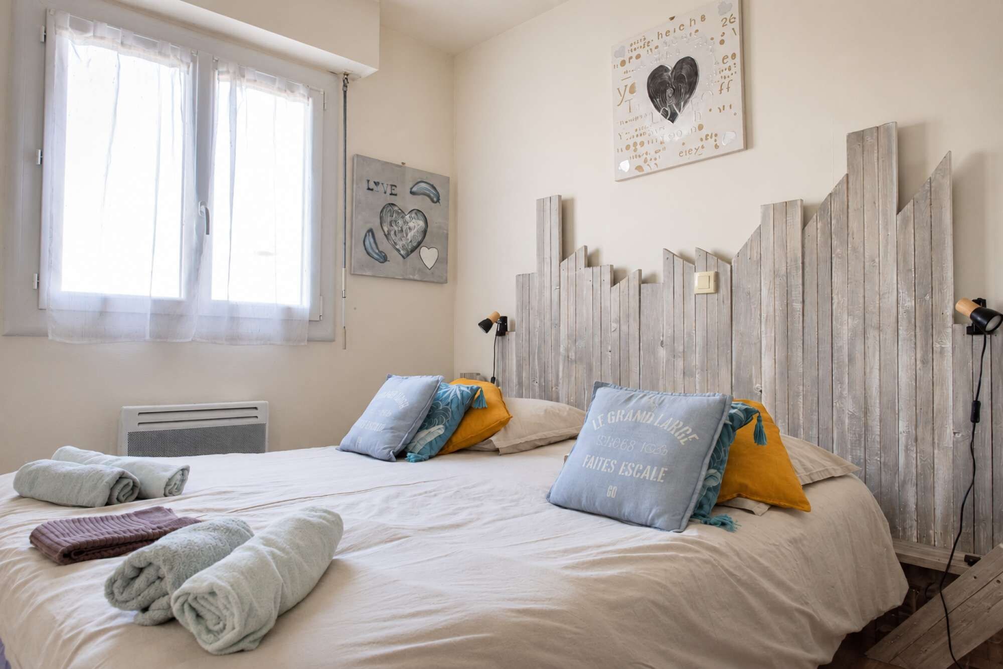 Un dormitorio con dos camas, una ventana con cortinas traslúcidas, una obra de arte en la pared y un cabecero de madera.