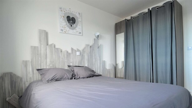 A bedroom with a bed, a headboard with vertical wooden slats, a wall-mounted light fixture, and a window with curtains.