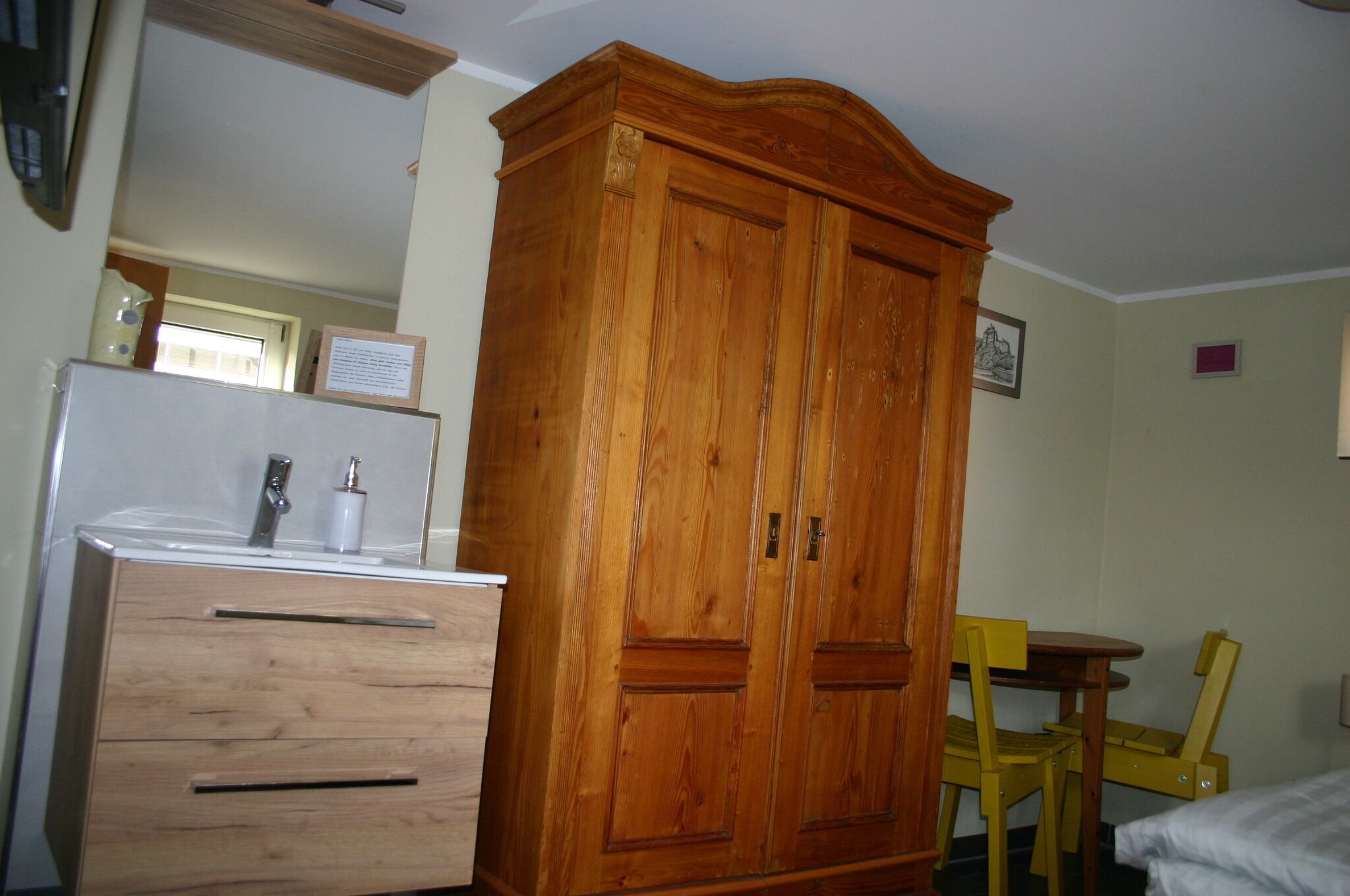 A wooden wardrobe with two doors, a wooden dresser with drawers, a small round table with two chairs, and a bed with white bedding.