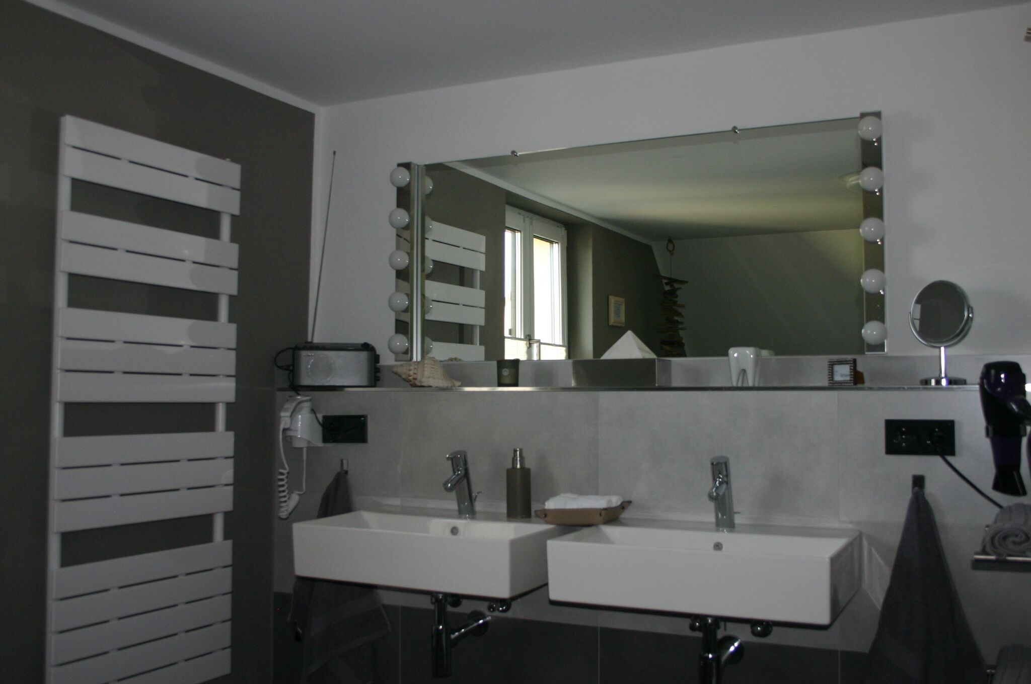 Een moderne badkamer met twee wastafels, een grote spiegel met verlichting, een handdoekrek en een haardroger.