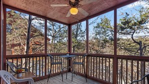 Une terrasse extérieure couverte, dotée d’une table et de chaises, d  ’un ventilateur de plafond, et offrant une vue sur des arbres et un bâtiment en arrière-plan.