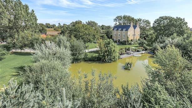 Un grand bâtiment avec un étang au premier plan, entouré d’une végétation luxuriante et d’arbres.