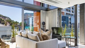 A modern living room with a sofa, TV, and glass table, large windows with a city view, and a balcony with plants. - Always-Power Stunning Apartment with 180 Degree City View near V&A Waterfront (Cape Town)