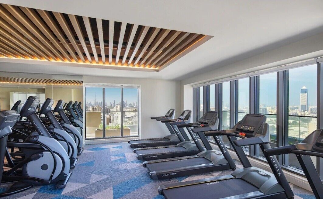 A gym with treadmills and exercise equipment, large windows with a city view, and a modern ceiling design.