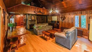 A cozy cabin interior with a wooden ceiling, a fireplace, a sofa, a dining table, and a large window. - The Pebblestone Cottage - Quaint Countryside Escape (Farmington)