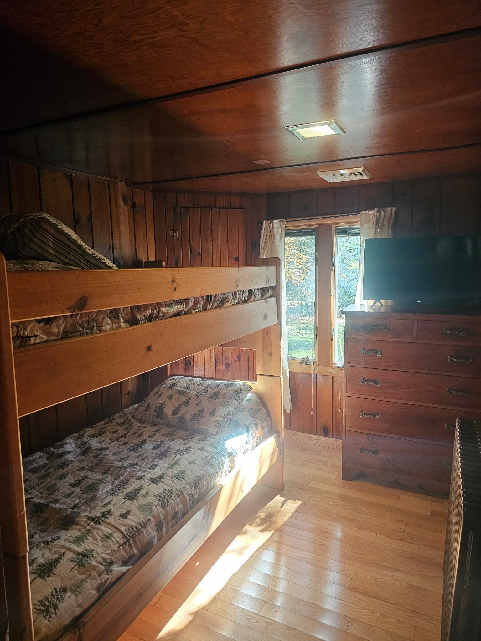 A bunk bed room with a wooden floor, a dresser, and a window with curtains.