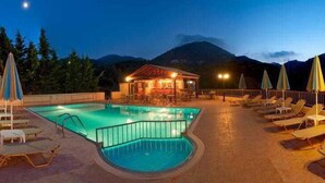 A swimming pool with lounge chairs and umbrellas, a small building with a red roof, and a mountain in the background. - Elpis Studios And Apartments (Mylopotamos)