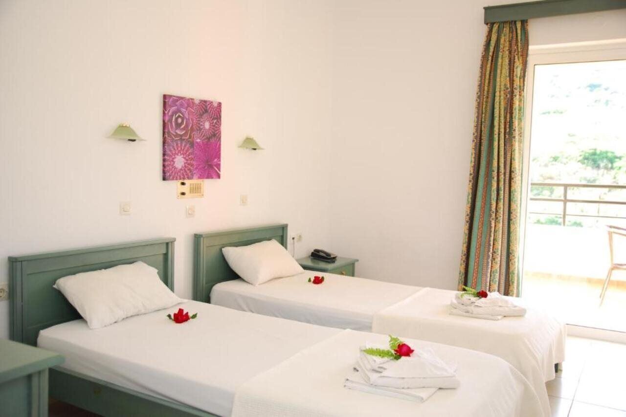 Habitación de hotel con dos camas individuales, una mesita, una lámpara de pared y un ventanal con cortinas.