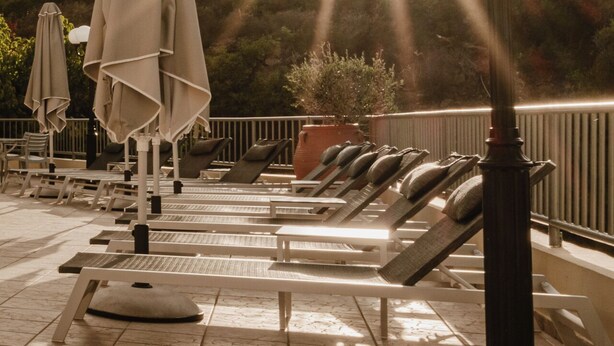 A sunlit outdoor seating area with lounge chairs and umbrellas, overlooking a mountainous landscape. - Elpis Studios And Apartments (Mylopotamos)