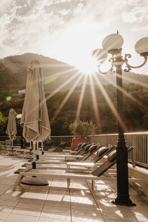 A sunlit outdoor seating area with lounge chairs and umbrellas, overlooking a mountainous landscape. - Elpis Studios And Apartments (Mylopotamos)