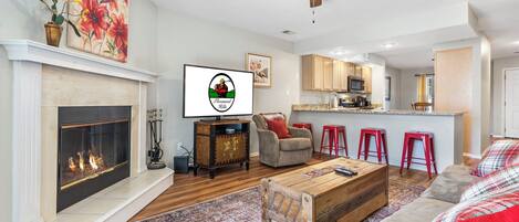 A living room with a fireplace, a ceiling fan, a kitchen in the background, and a red bar stool.