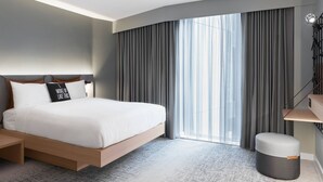 A modern hotel room with a large bed, a bedside table, a small ottoman, and a view of the city through large windows. - Moxy Slough (Slough)