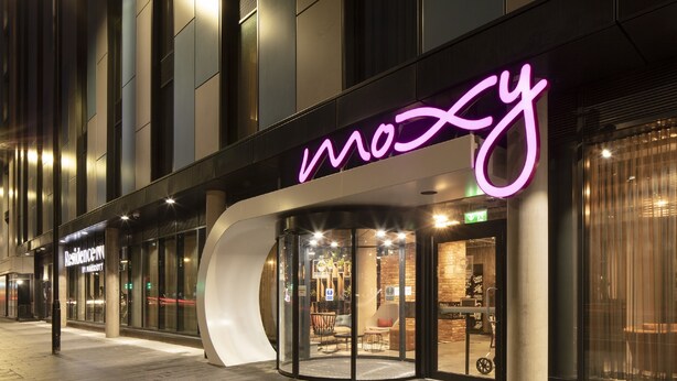 A modern hotel entrance with a neon sign, glass doors, and a seating area visible inside. - Moxy Slough (Slough)