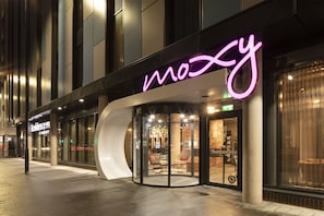 A modern hotel entrance with a neon sign, glass doors, and a seating area visible inside.