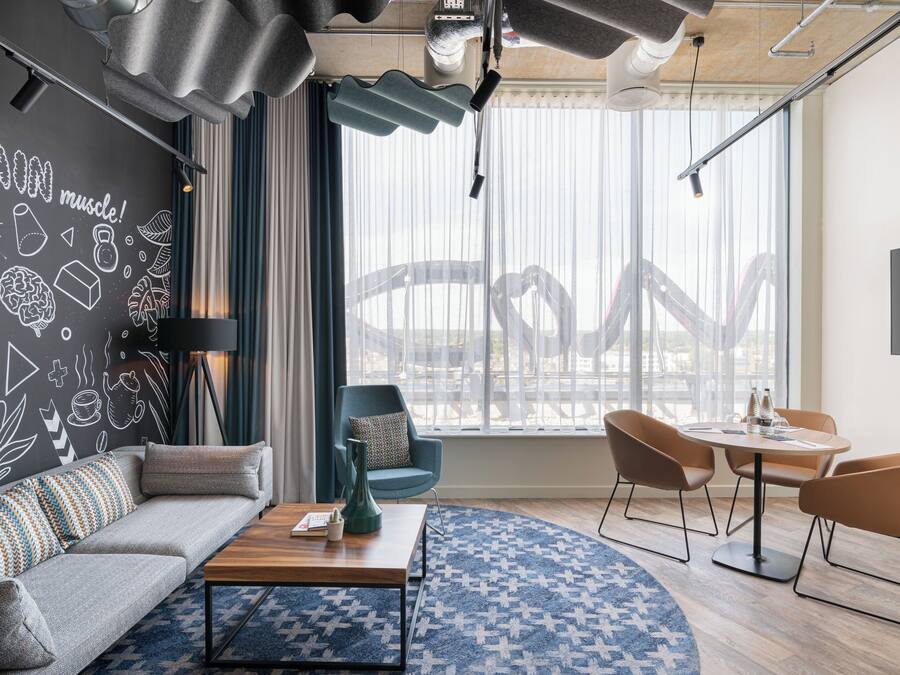 Una sala de estar moderna con sofá, mesa de centro y comedor con sillas. Hay un ventanal con persianas, un televisor montado en la pared y una pared decorativa con formas y texto variados.