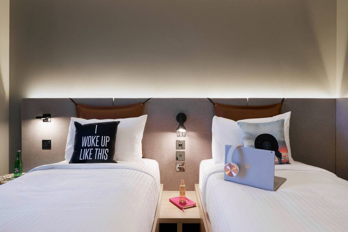 A hotel room with two beds, each with a pillow, a bedside table with a laptop, a bottle, and a notebook.