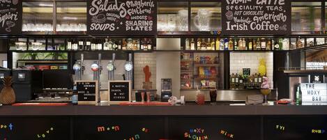 Zona de bar con una pizarra que muestra un menú con hamburguesas, nachos y café con leche. Hay taburetes y una barra con diversas bebidas y aderezos.