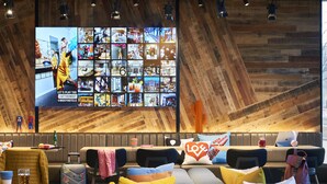 A modern lounge area with seating, a large screen displaying various images, and a wooden wall. - Moxy Slough (Slough)
