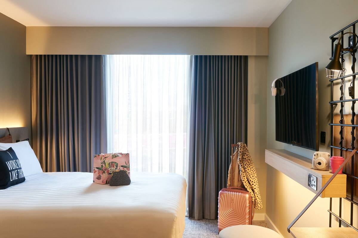 A hotel room with a large bed, a flat-screen TV mounted on the wall, a desk with a lamp, and a suitcase.