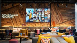 A modern hotel lobby with a large TV displaying a 'RECORDING IN PROGRESS' sign, comfortable seating areas, and a wooden accent wall. - Moxy Slough (Slough)