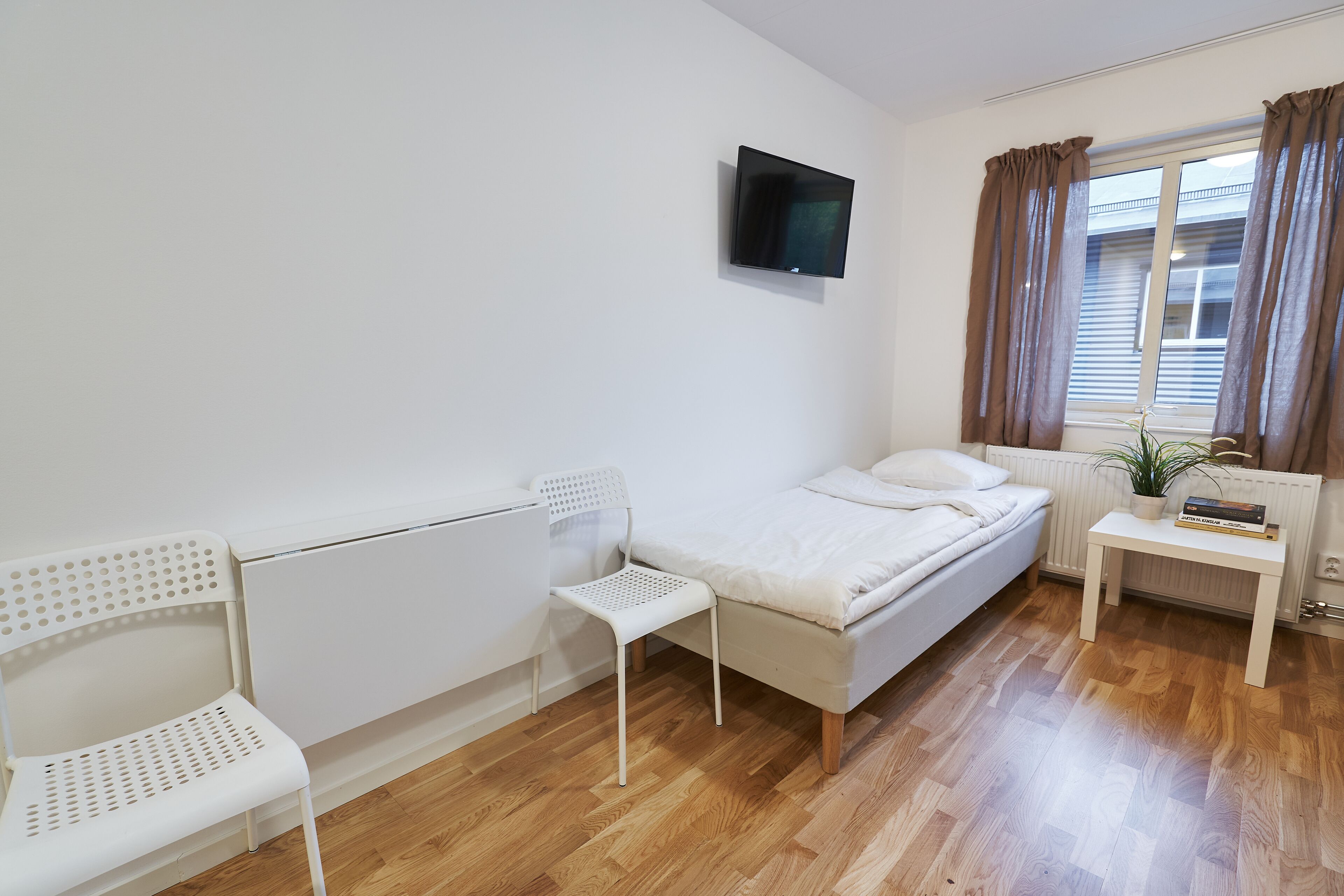 A small, clean room with a bed, a chair, a TV, and a window with curtains.