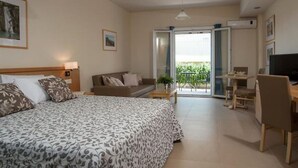 A hotel room with a bed, a sofa, a desk, and a TV. There is a balcony with a view of greenery. - Estia Hotel (Pylos-Nestoras)